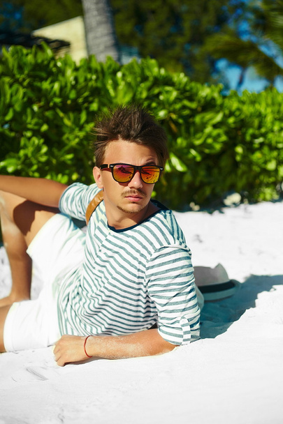 Stylish young male model man lying on beach sand  wearing hipster summer hat  enjoying summer travel holiday near ocean in sunglasses - Photo, Image