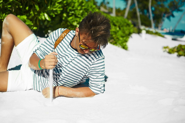 Stylish young male model man lying on beach sand  wearing hipster summer hat  enjoying summer travel holiday near ocean in sunglasses - Foto, Imagen
