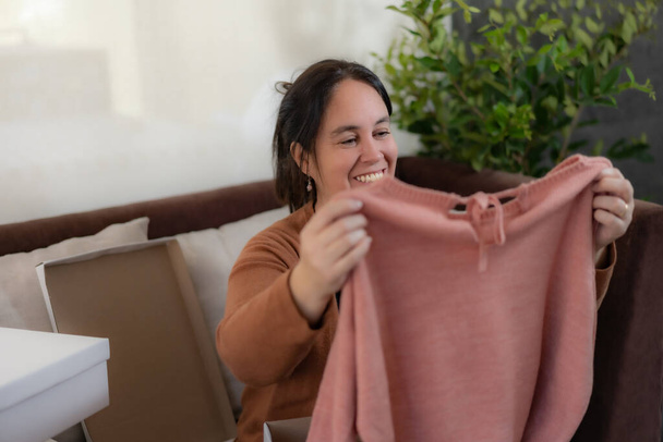 a very happy lady because she received a gift in a white box on a special day. She opens the gift box and inside is a pink sweater. She takes it out of the box and looks at it. - Foto, immagini