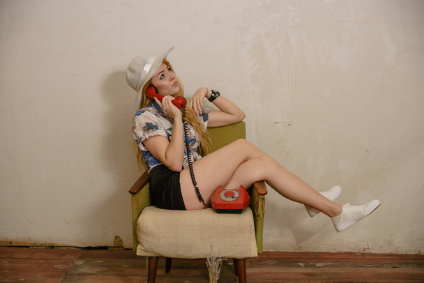 красивая блондинка сидит в кресле с красным винтажным телефоном - Фото, изображение