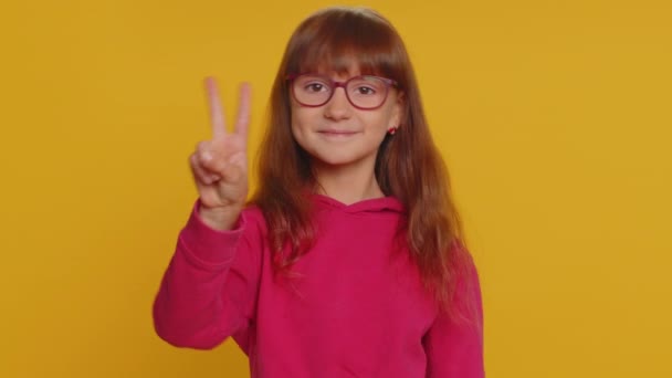 Happy preteen child girl kid showing victory sign, hoping for success and win, doing peace gesture smiling with kind optimistic expression. Little toddler children isolated on studio yellow background - Footage, Video