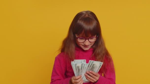 Gazdag elégedett fiatal kisiskolás lány szemüvegben integetett pénzt dollár bankjegyek számlák, mint egy rajongó, siker, lottó nyertes, nagy jövedelem, gazdagság. tini női gyermek gyerek a stúdióban sárga háttér - Felvétel, videó