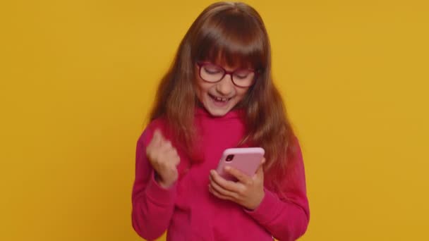 Young preteen child girl kid use mobile smartphone typing browsing say Wow yes found out great big win good news celebrate doing winner gesture. Little toddler children on studio yellow background - 映像、動画