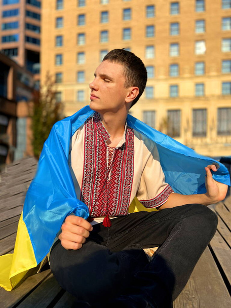 Un jeune homme assis dans une chemise brodée sur ses épaules avec le drapeau de l'Ukraine En arrière-plan est grand bâtiment jaune avec des fenêtres Il a tourné son visage au soleil symboles ukrainiens terrorisme russe 2022 - Photo, image