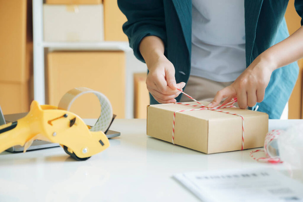 Female small business entrepreneur, online store owner packing and tying parcel with rope ready for sending to post office for shipping to customer. Online selling, e-commerce cencept. - Foto, afbeelding