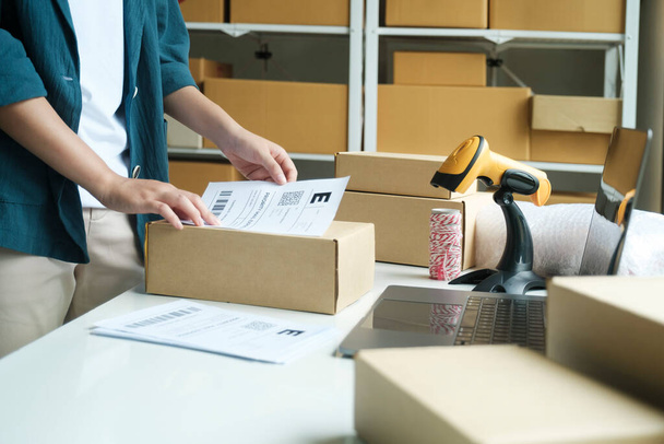 Young small business entrepreneur woman, online store onwer with shelves of cardboard boxes in the background packing product in mailing box, labeling delivery package for shipping to customer. Online - Fotoğraf, Görsel
