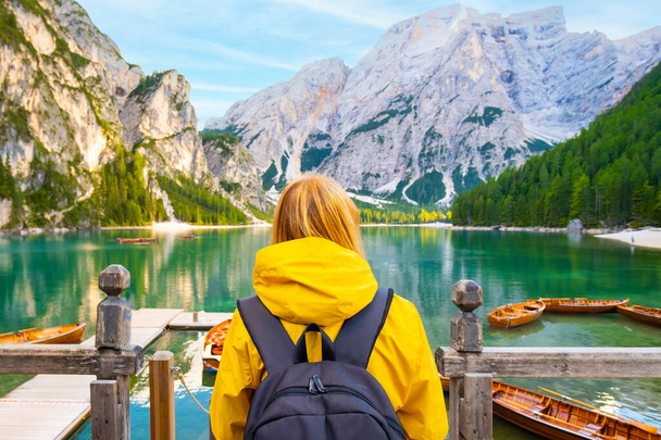 バックパック付きの女性の背景とドロマイトでターコイズブルーの水と木製のボートを持つ湖で楽しい黄色のスポーツジャケットを身に着けています. - 写真・画像