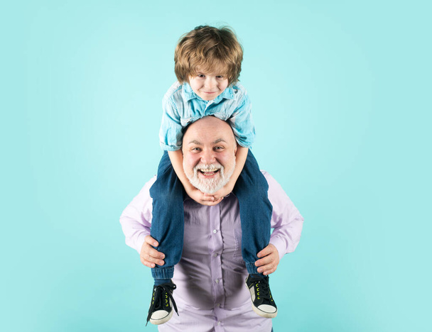 Grandfather and son piggyback ride with funny face isolated on blue in studio. Granddad and cute boy with funny face - Zdjęcie, obraz