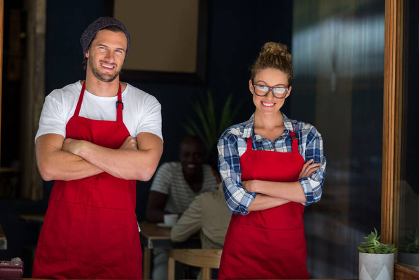 Улыбающаяся официантка и официант, стоящие со скрещенными руками возле кафе - Фото, изображение