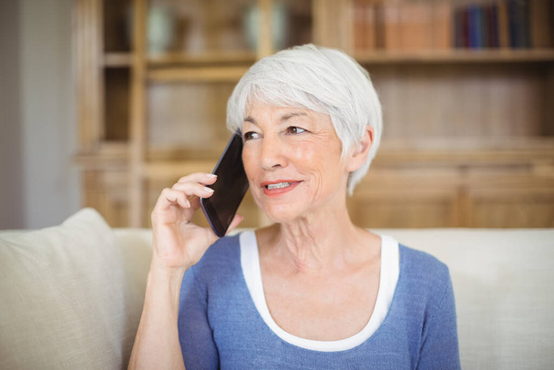 Hymyilevä vanhempi nainen puhuu matkapuhelimella olohuoneessa
 - Valokuva, kuva