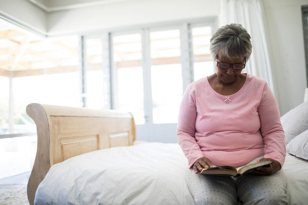 Oudere vrouw die boek leest terwijl ze op bed zit in de slaapkamer - Foto, afbeelding