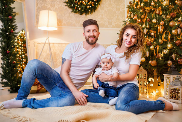 Happy family of mother, father and little baby girl sitting near Christmas tree on the floor in decorated interior. Celebrating Christmas.  - Fotoğraf, Görsel
