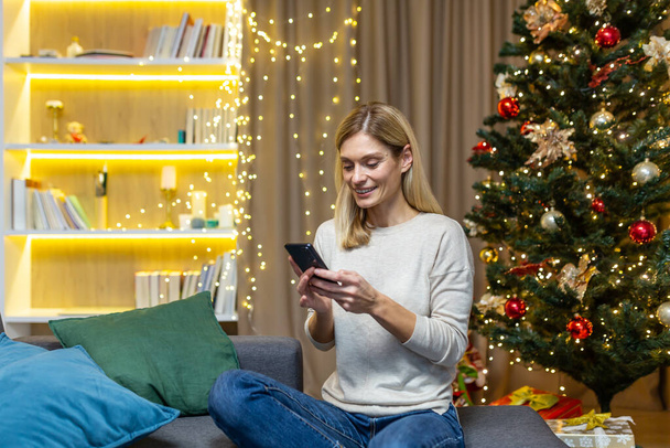 A happy young woman is holding a phone and placing an order for food delivery for a festive dinner. Sitting on sofa at home with New Year and Christmas decorations, lights and Christmas tree. - Fotó, kép