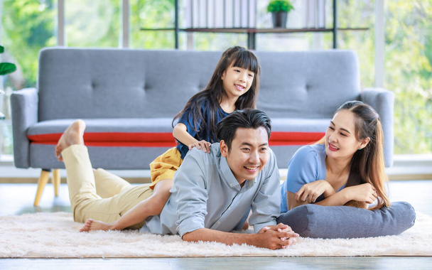 千年のアジアの素敵な幸せな家族のお父さんのお母さんはカーペットの床に横たわっています。若い娘女の子座っていますpiggyバックオンお父さん笑顔一緒に笑って再生で自宅で週末. - 写真・画像