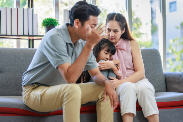 Millennial Asian unhappy family mother sitting on sofa couch holding hugging soothing little young female daughter while sad stressed father crying after big fight argument in living room at home. - Photo, Image