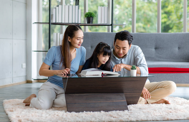 Millennial Asian happy family father and mother sitting on cozy carpet floor smiling helping teaching little young girl daughter reading learning with big textbook in living room at home. - Фото, изображение