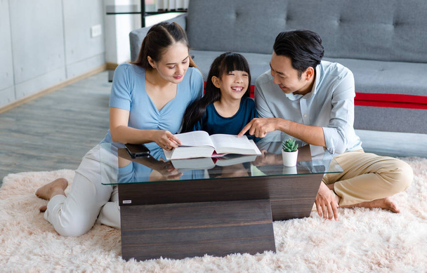 Millennial Ασίας ευτυχισμένη οικογένεια πατέρας και μητέρα κάθεται στο ζεστό πάτωμα χαλί χαμογελώντας βοηθώντας τη διδασκαλία κοριτσάκι κόρη ανάγνωση σπουδάζοντας μάθηση με μεγάλο βιβλίο στο σαλόνι στο σπίτι. - Φωτογραφία, εικόνα