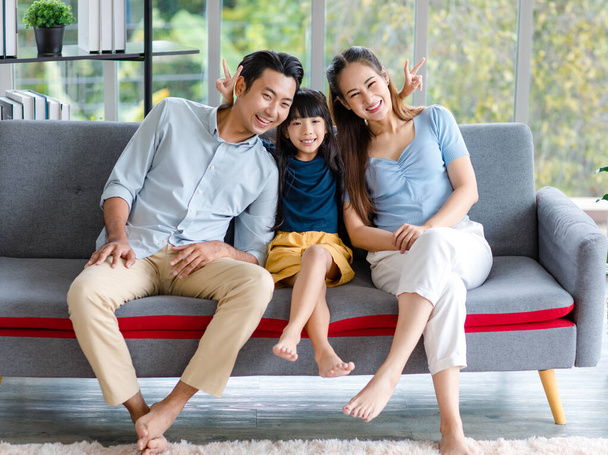 Millennial asiático feliz familia padre madre y pequeño niño pequeño niña sentado sonriendo mirando a la cámara juntos en el sofá acogedor sofá que muestra dos dedos signo de paz tomar fotos en la sala de estar en casa. - Foto, imagen
