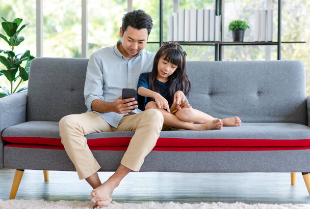 Millennial Asian happy family father sitting smiling on cozy sofa couch holding showing funny content streaming online via touchscreen smartphone to little young girl daughter in living room at home. - Photo, Image