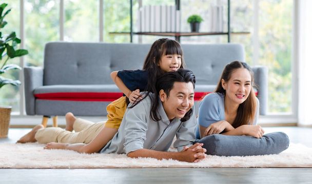 Millennial Asian lovely happy family father mother laying down on carpet floor while young daughter girl sitting piggy back on dad smiling laughing playing together in living room at home in weekend. - Фото, изображение