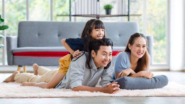 Millennial Azji piękny szczęśliwy rodzina ojciec matka leżąc na podłodze dywan podczas gdy młoda córka dziewczyna siedzi świnka z powrotem na tata uśmiechnięty śmiech bawiąc się razem w salonie w domu w weekend. - Zdjęcie, obraz