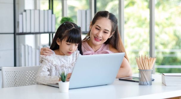 Millennial Azjatycki szczęśliwy rodzina matka uśmiech pomaga wspieranie nauczania dziewczynka córka studiuje naukę odrabiania lekcji w szkole online za pośrednictwem laptopa notebook komputer w salonie w domu. - Zdjęcie, obraz