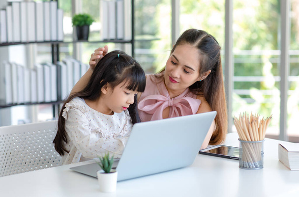 Millennial Asian happy family mother smiling helping supporting teaching little girl kid daughter studying doing online school homework via laptop book computer in living room at home. - Фото, изображение