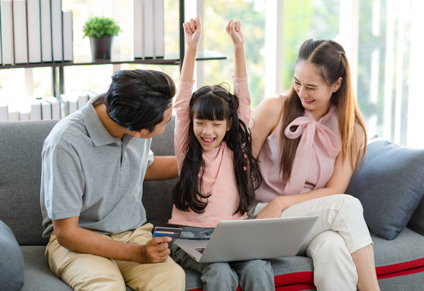 Azji szczęśliwy podekscytowany dziewczynka córka siedzi na przytulnej kanapie uśmiech krzyki podczas gdy ojciec i matka za pomocą karty kredytowej dokonywania płatności zakupy prezent obecny online za pośrednictwem laptopa. - Zdjęcie, obraz