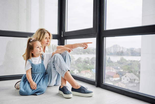 Γυναίκα και κοριτσάκι κάθονται στο πάτωμα και κοιτάζουν και δείχνουν έξω από το παράθυρο στο νέο οικογενειακό σπίτι. Μητέρα και παιδί απολαμβάνουν θέα από πανοραμικό παράθυρο σε νεόκτιστο διαμέρισμα. - Φωτογραφία, εικόνα