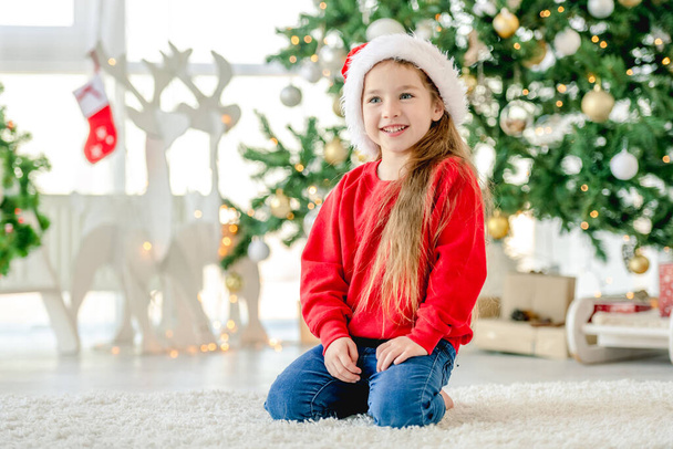Noel zamanı Noel Baba şapkası takan küçük bir kız Noel ağacıyla süslenmiş evinde oturuyor ve gülümsüyor. Noel zamanı tatlı bir çocuk. - Fotoğraf, Görsel