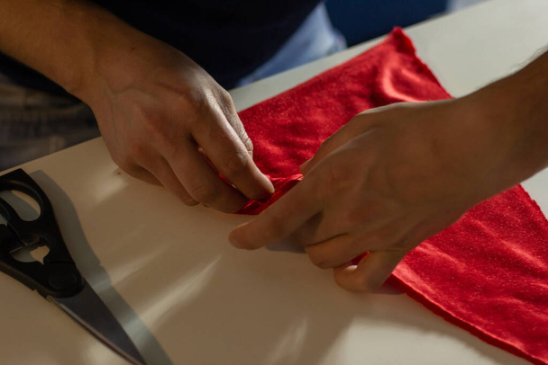 Hands of experienced worker in the handmade industry, performing cutting tasks on red fabric, on a white surface, the beginning of red hat production. Homemade sewing. Working tailor. Christmas hat. - Foto, Imagen