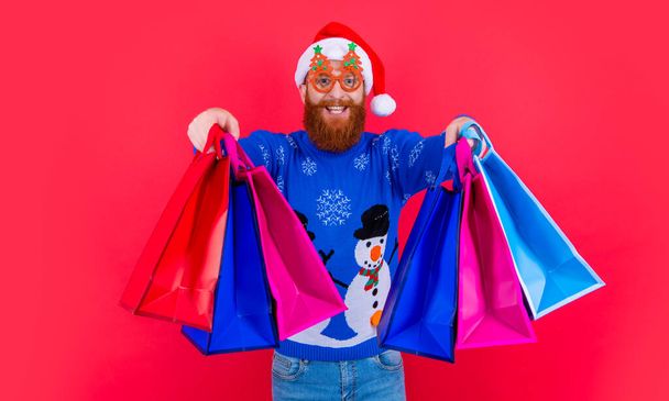 xmas shopping sale. cheerful man with shopping bag after xmas sale. santa man with xmas shopping bag at sale isolated on red. - Zdjęcie, obraz