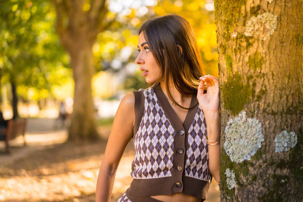 Brunette model in autumn at sunset in a natural city park, next to a tree - Φωτογραφία, εικόνα