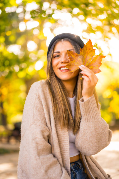 Young blonde girl with a wool cap in autumn, with a leaf on her face in the sunset smiling - 写真・画像
