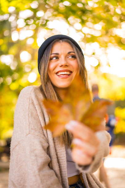 Junge blonde Mädchen mit einer Wollmütze im Herbst, mit einem Blatt auf dem Gesicht im Sonnenuntergang lächelnd - Foto, Bild