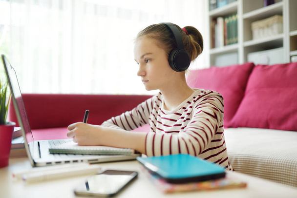  Egy kamasz lány, aki a házi feladatát csinálja laptoppal otthon. A gyerek szerkentyűket használ a tanuláshoz. Online oktatás és távoktatás gyerekeknek. Magániskola a karantén ideje alatt.  - Fotó, kép