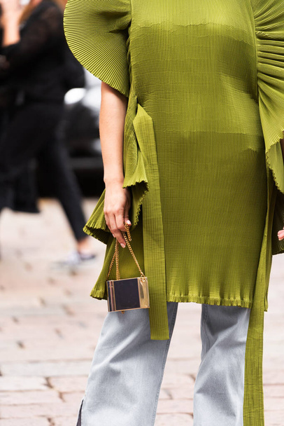 Mediolan, Włochy - 21 września 2022: Kobieta nosi khaki zieloną krótką sukienkę z przerośniętym akordeonem falistą krótką spódniczkę, srebrną metaliczną małą torebkę od Diora, niebieskie, wyblakłe spodnie typu fllared - Zdjęcie, obraz