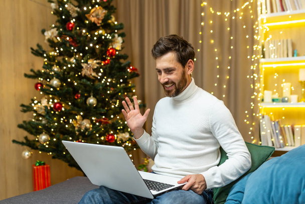 Man at home recording video greetings, Merry Christmas and Happy New Year, sitting on sofa at home near Christmas tree, using laptop for online remote communication with friends, video call. - Fotoğraf, Görsel