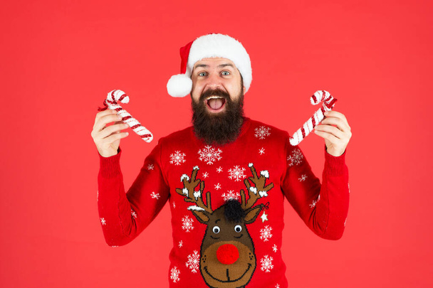 brutal hipster in winter knitted xmas sweater. mature guy with holiday candy stick on red background. happy new year. merry christmas. amazed bearded man in santa claus hat. - Fotó, kép