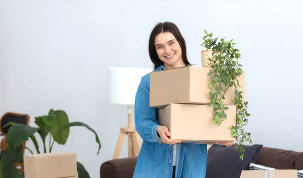 Smiling pretty caucasian woman standing in her new flat with a stack of cardboard boxes and friendly smiling looking at the camera. Moving to a new house, rent - Фото, изображение