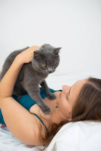 Νεαρή γυναίκα κρατάει απαλά τη Βρετανίδα γάτα της στο κρεβάτι της - Φωτογραφία, εικόνα