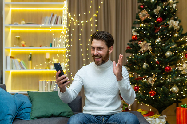 A man talks on a video call at home with a phone, sits on a sofa near a Christmas tree, records video greetings on New Years and Christmas holidays. - 写真・画像