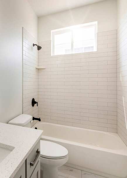 Vertical Sun flare Salle de bain avec fenêtre, intérieur blanc et luminaires noirs. Il y a un lavabo sur la droite avec dessus en granit et miroir près des toilettes à côté de la baignoire avec pomme de douche murale. - Photo, image