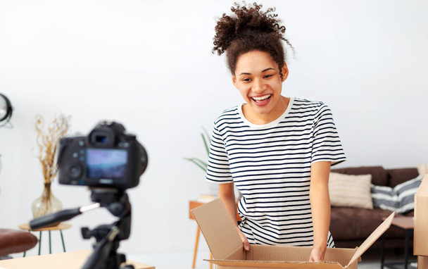 Young woman unpacking boxes on camera. African American female small business owner, blogger vlogger freelancer using camera to recording video content for his followers - 写真・画像