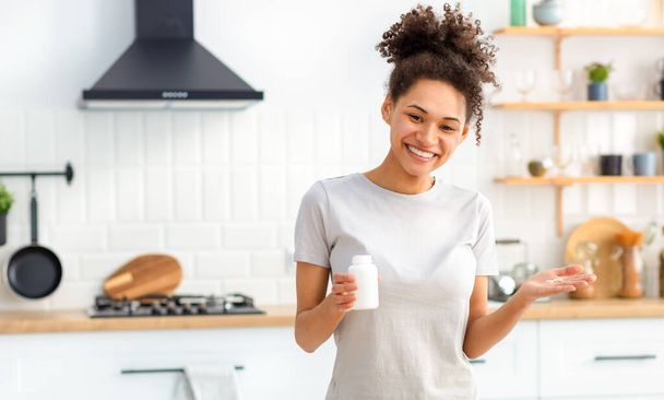 Felice donna afroamericana in cucina in casa con una bottiglia di integratori alimentari, guardando la fotocamera e sorridente amichevole, stile di vita sano - Foto, immagini
