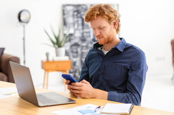 Concentrated man entrepreneur freelancer sitting in modern office chatting using smartphone Young businessman, small business owner male working, writes message using mobile phone discussing something - 写真・画像
