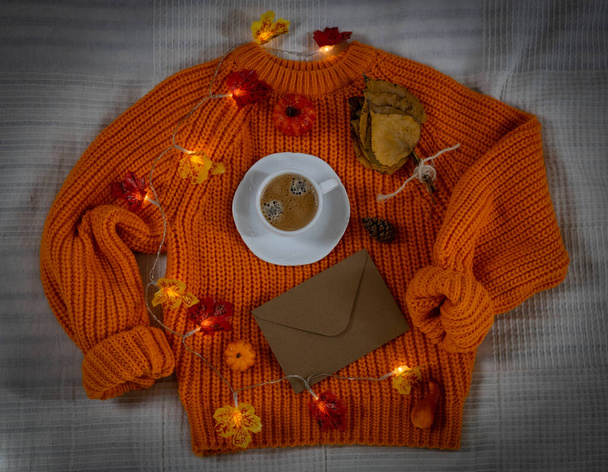 Una piccola tazza di caffè con un piattino, una ghirlanda ardente di foglie autunnali, coni, piccole zucche, una busta vuota artigianale e un mazzo di foglie di pioppo su un maglione a maglia arancione, vista dall'alto. - Foto, immagini