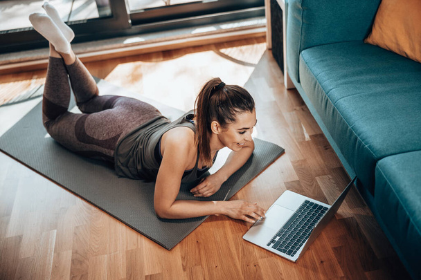 Молодая женщина с помощью ноутбука во время занятий фитнесом дома. - Фото, изображение