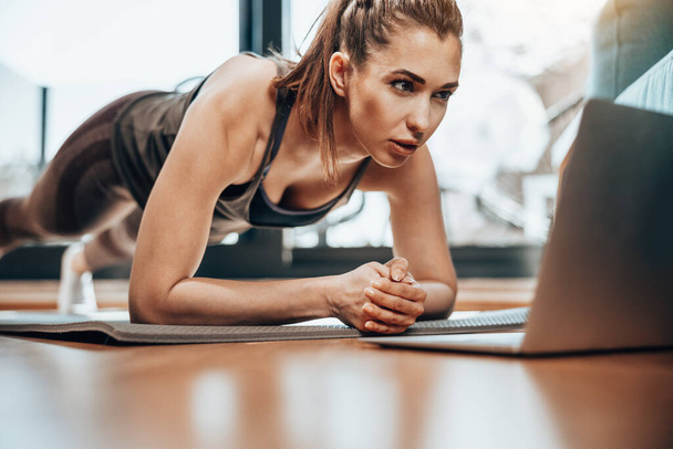 Young woman using laptop while doing plank exercises at home. - Фото, изображение
