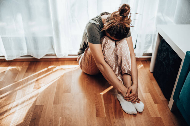 Eenzame jonge vrouw in depressie zitten door het raam met omhelzing knieën verloren in droevige gedachten. - Foto, afbeelding
